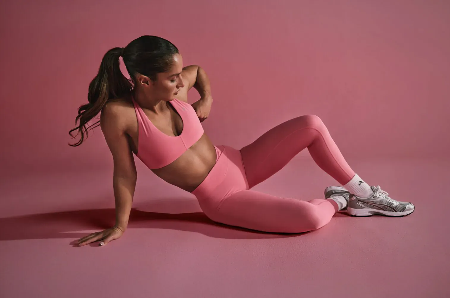 Fit woman in pink sports bra and leggings poses seated on pink background, ponytail and white sneakers.