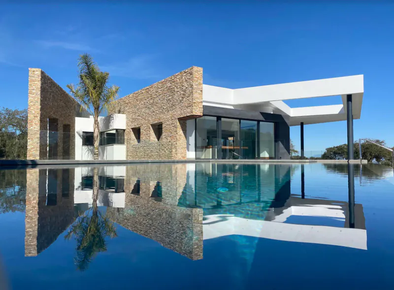 Modern stone and white villa with infinity pool reflecting in blue water, palm tree, blue sky, Spain.
