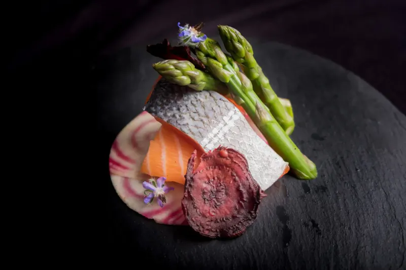 Artistic plate of salmon sashimi with asparagus spears, beet slices, and edible flowers on black slate