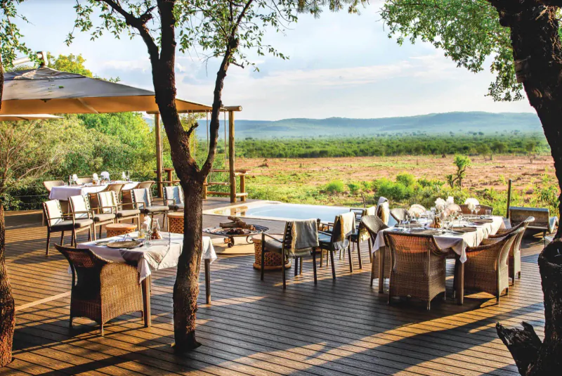 Elegant outdoor dining deck at Madikwe Hills lodge with tables set under umbrellas, acacia trees, savanna views.