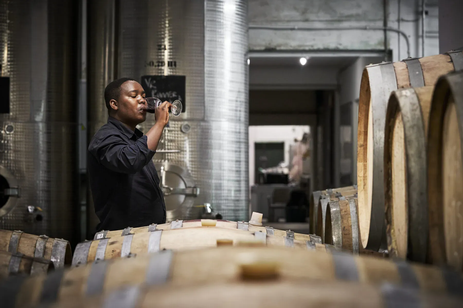 Black man in dark shirt sips red wine from glass amid oak barrels and stainless steel tanks in winery