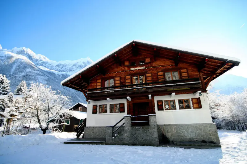 Wooden chalet with snow-covered roof and balcony in snowy alpine mountains under blue sky