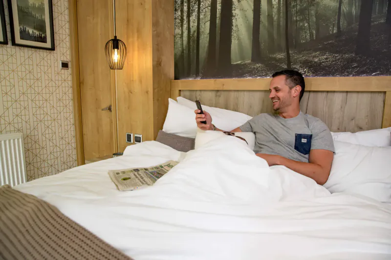 Man in grey t-shirt sits up in white-sheeted bed in New Forest-themed hotel room, using phone and reading newspaper