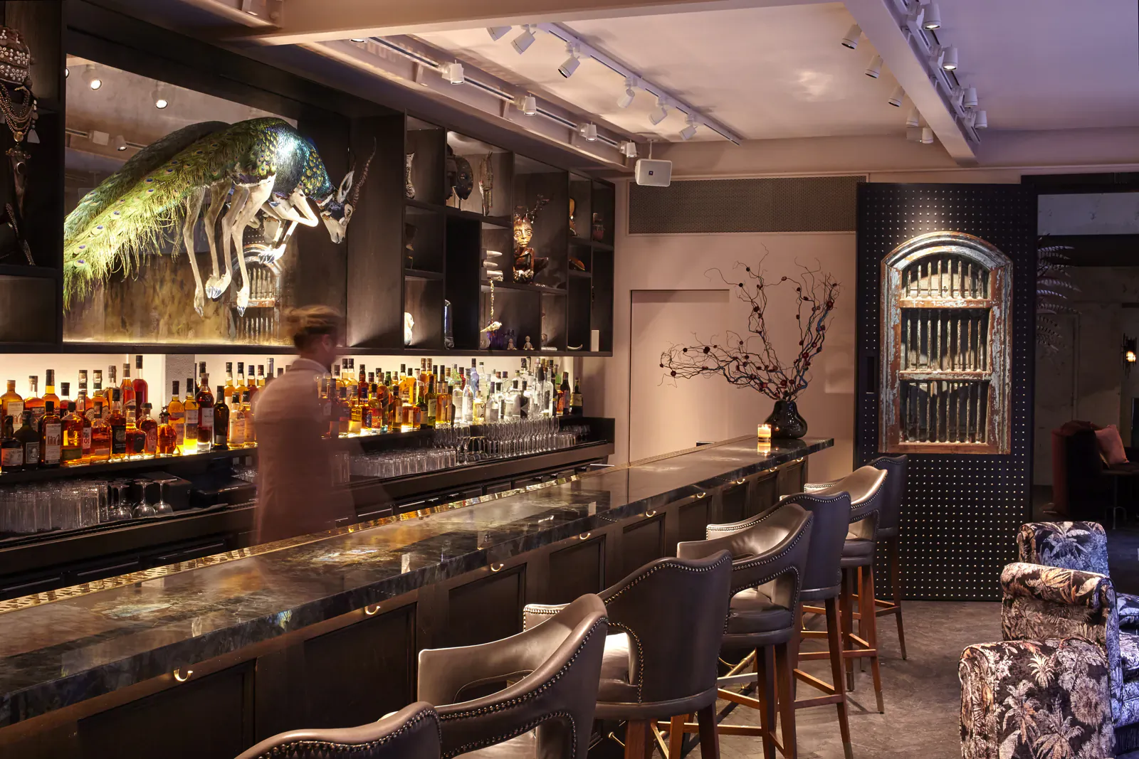Modern hotel bar with backlit giant insect artwork, extensive liquor shelves, bartender behind marble counter, and blue leather barstools.