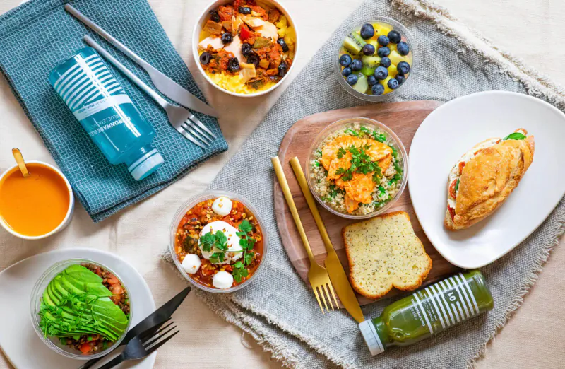 Cojean Selfridges overhead view of healthy bowls, soups, sandwich, bread, berries, and drinks on table with cutlery.