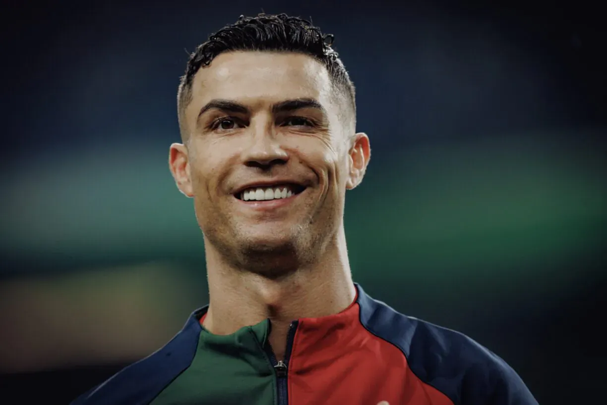 Cristiano Ronaldo smiles confidently in red, green, navy jacket on stadium field