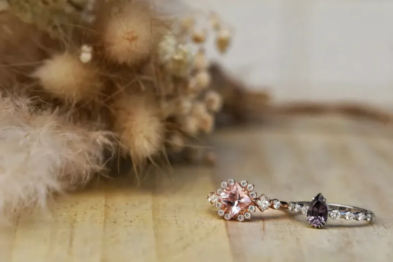 Pink flower-cut diamond ring and pear-shaped purple gem ring on wooden surface with dried pampas grass backdrop.