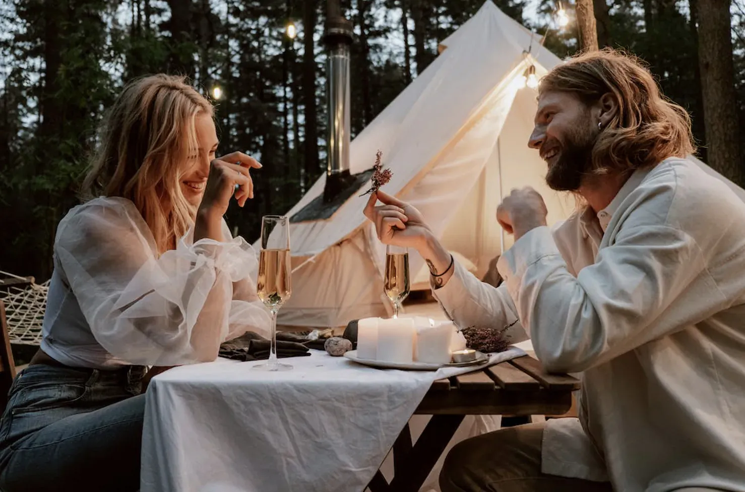 Smiling couple enjoying romantic candlelit dinner outside white tent in forest at dusk with wine glasses and cake
