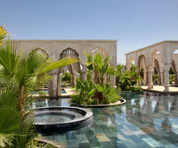 Moroccan-style courtyard with arched stone colonnades, palm trees, and reflective pool under blue sky
