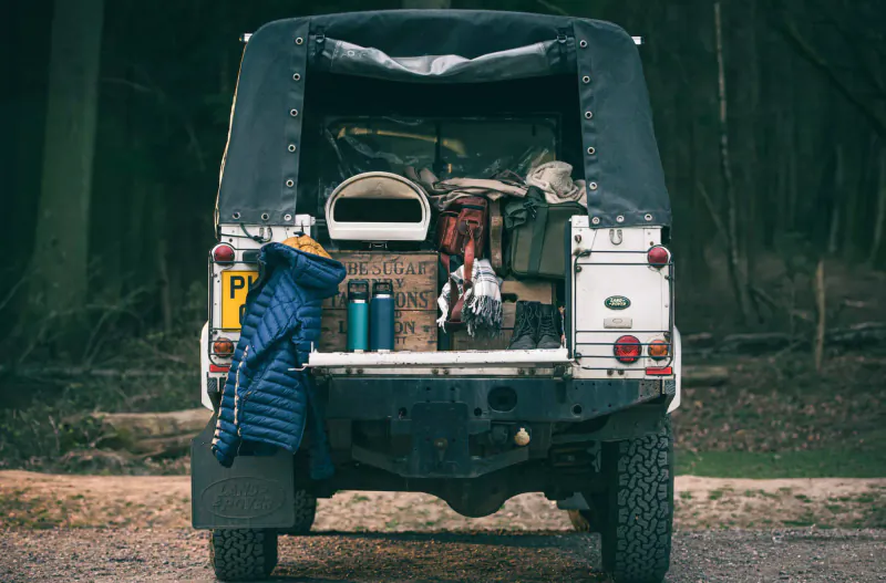 Rear of white Land Rover Defender packed with camping gear like jacket, boxes, stove in forest setting
