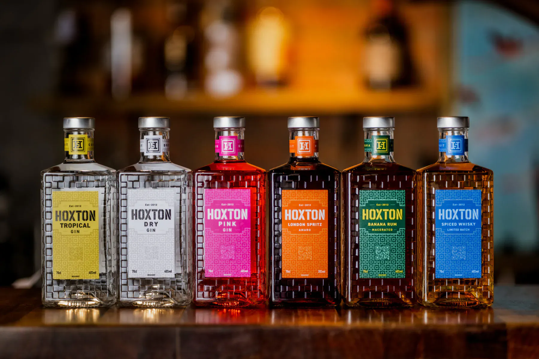 Six colorful Hoxton Spirits bottles in new design on bar counter with blurred bar background.