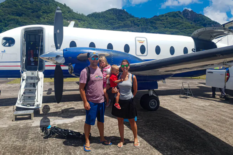 Family with toddler posing in front of white prop plane on Alphonse Island airstrip, lush mountains and blue sky backdrop.