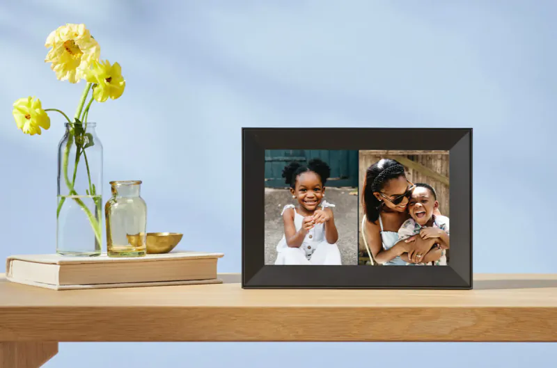 Black Aura smart frame on wooden table with two photos: smiling Black girl and mother holding baby boy, beside yellow flowers and vase.