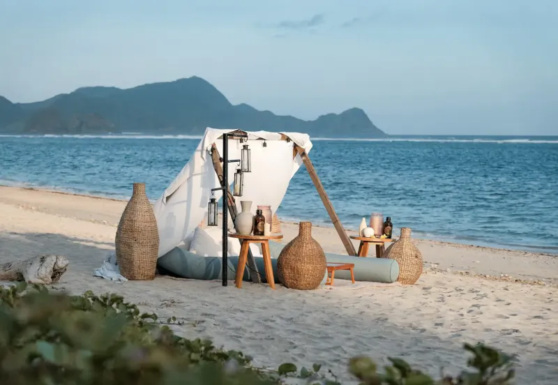 Romantic beach dining setup at Amber Lombok with white canopy, lanterns, bottles on tables, ocean and mountain views.