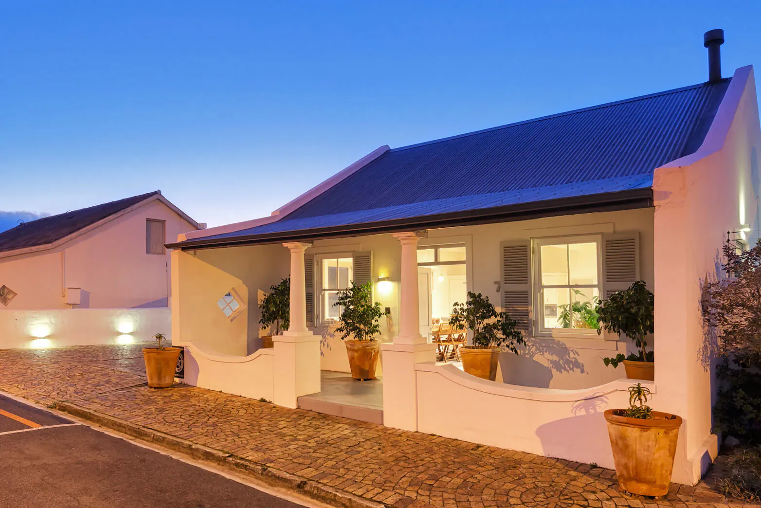 White Vishuis Cottage with blue roof, potted plants on veranda, lit warmly at dusk beside another cottage