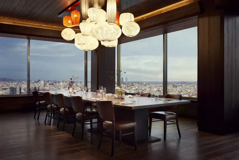 Elegant dining room in Nobu Hotel Barcelona with long wooden table, white and orange pendant lamps, city skyline view at dusk.