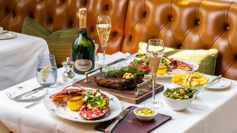 Elegant table at No. Fifty Cheyne with Veuve Clicquot champagne, grilled steak, lobster, fries, and salads on white plates against orange leather booth.