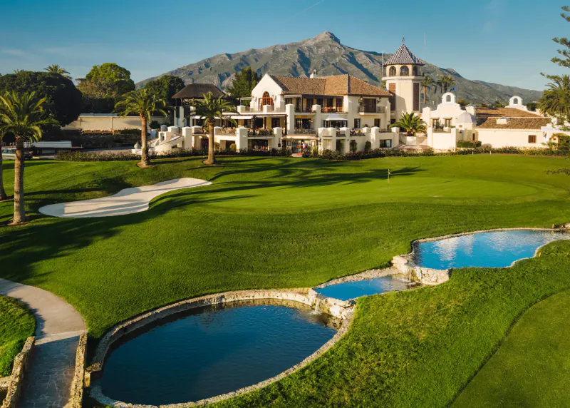 Aerial view of Los Naranjos Golf Club: luxurious white clubhouse with tower, lush green fairways, bunkers, ponds, palm trees, and Sierra Blanca mountains in Marbella.