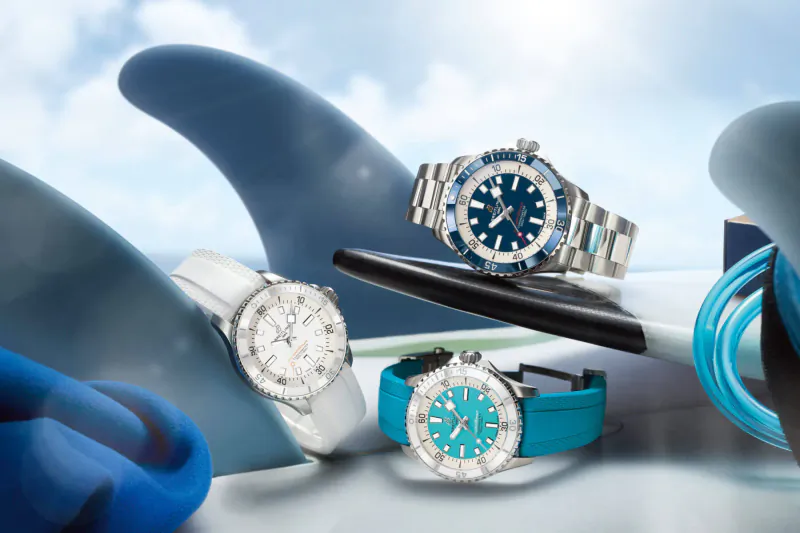 Three Breitling Superocean watches in blue and white on shark fin and surfboard setup, ocean theme.