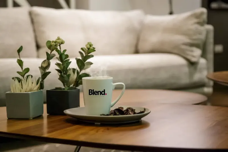 White 'Blend' mug steaming on saucer with cookies on wooden table beside plants and sofa