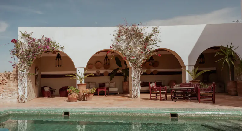 White arched Moroccan riad with hanging lanterns, plants, seating areas, and turquoise pool at Les Jardins De Villa Maroc in Essaouira