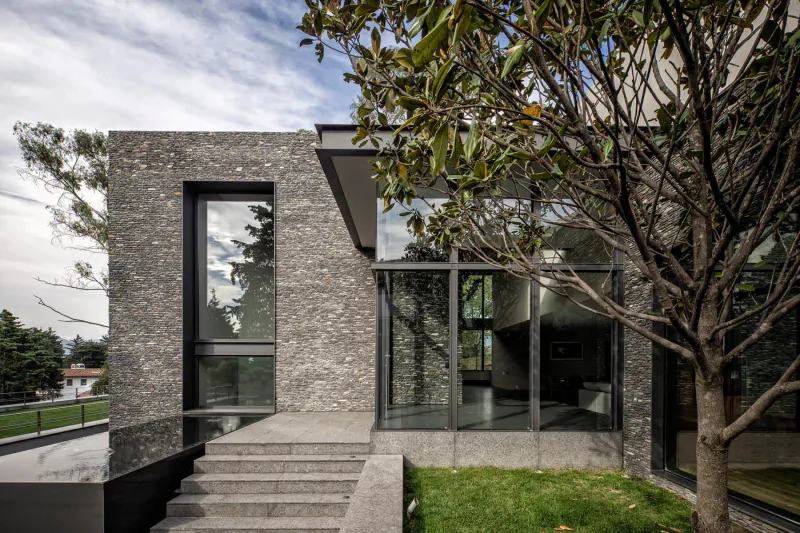 Modern Mexican stone house facade with large glass windows, concrete steps, grass, and overhanging tree under cloudy sky.