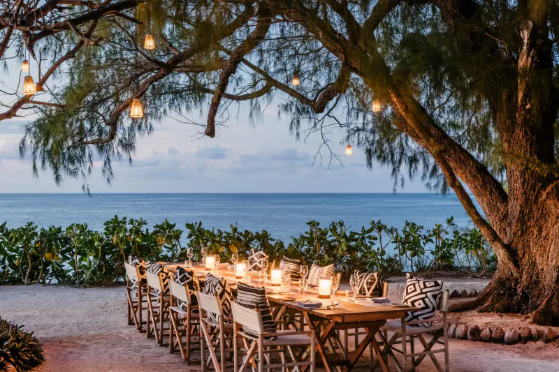 Elegant candlelit dinner table under string lights in large tree, beachfront at Four Seasons Desroches villa, sunset over ocean.