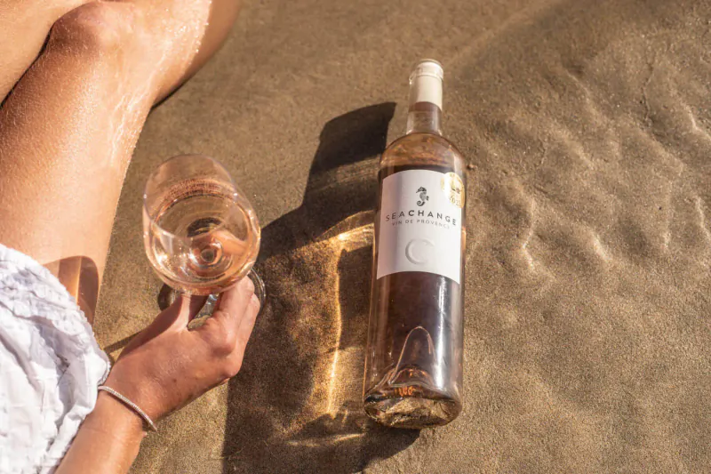 Woman's hand holding glass of rosé wine from 'Sea Change' bottle on sandy beach