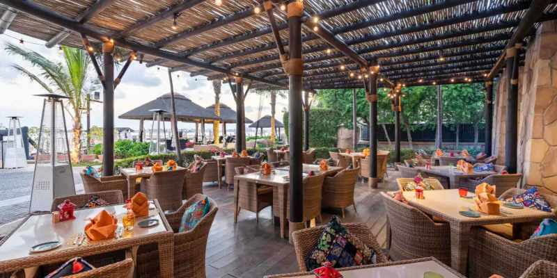 Open-air beachfront restaurant at Sofitel Dubai the Palm with thatched roof, wicker tables, colorful cushions, and tropical palms.