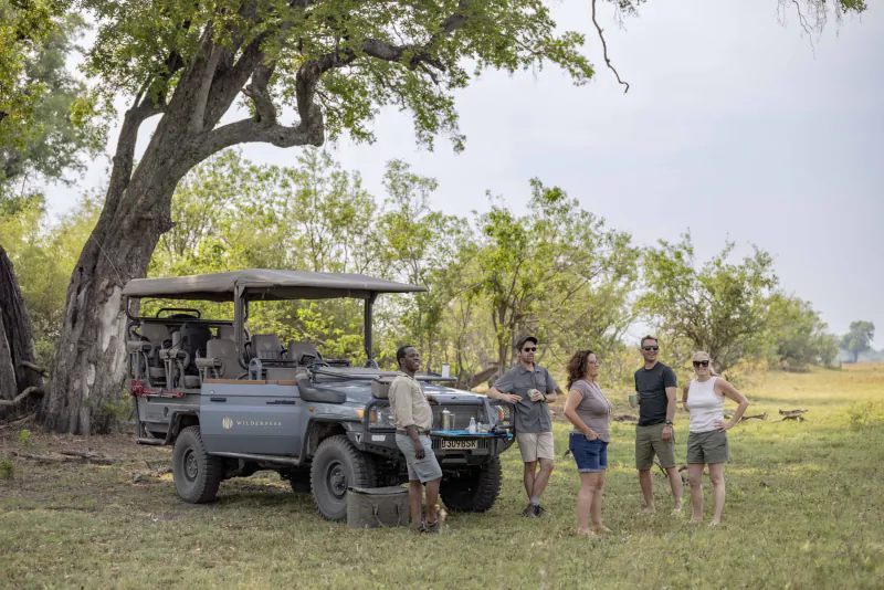 Group of four tourists in casual attire stand by gray safari jeep under large acacia tree in African savanna.