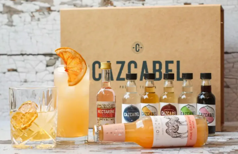 Open Cazzabel box with mini gin bottles, orange garnished highball and old fashioned cocktails on wood against brick wall