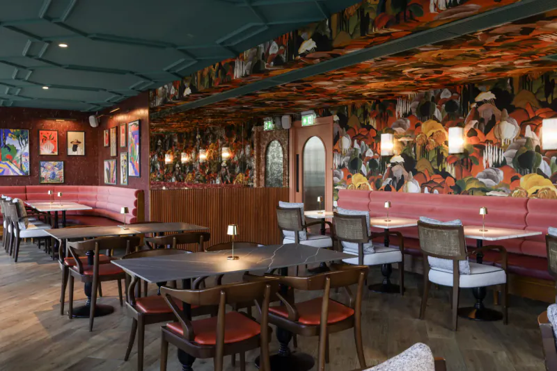 Luxurious restaurant interior with floral-patterned walls, curved red booths, wooden tables, and chairs under ornate green ceiling.