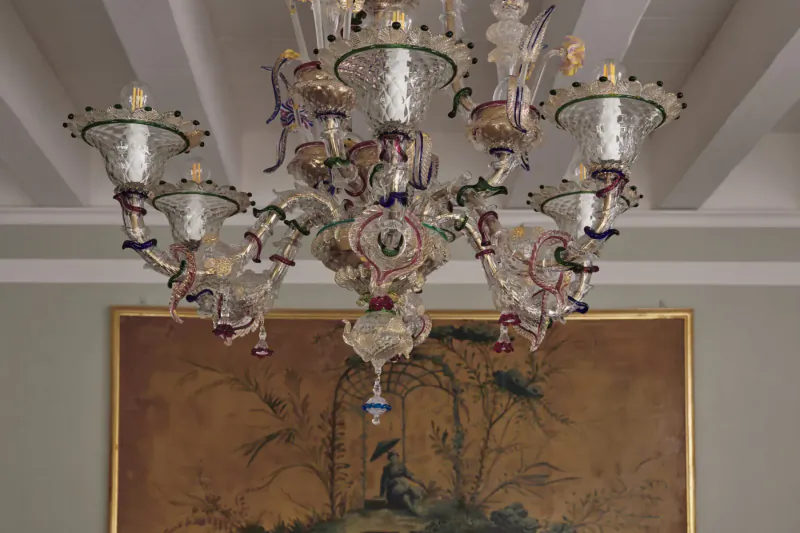Elegant multicolored Murano glass chandelier hanging from ceiling in front of gold-framed painting of trees and peacock in Venetian hotel.