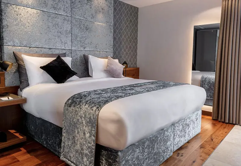 Luxurious king bed with gray velvet runner and pillows against textured wall in modern hotel room with mirror and wood floor.
