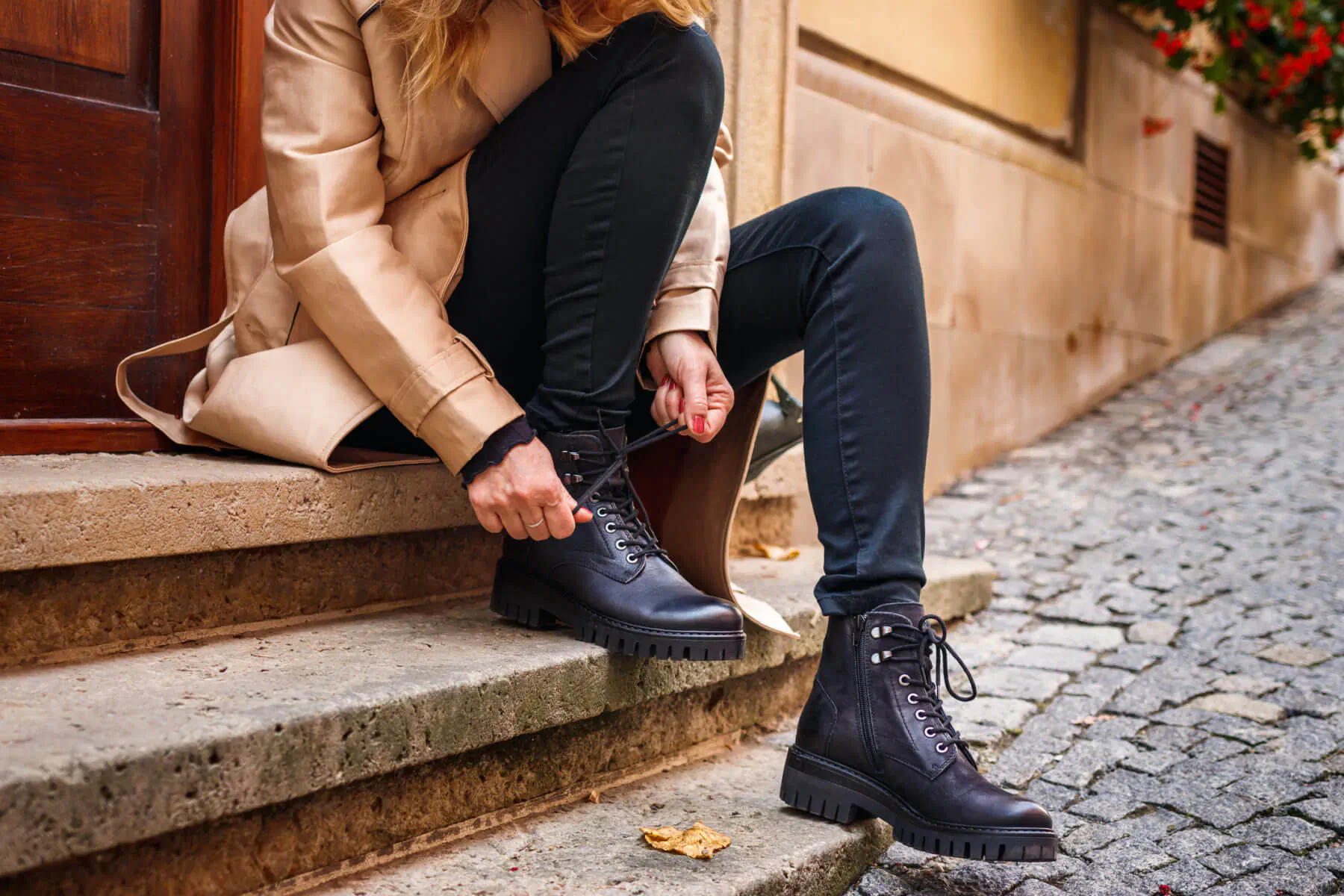 Woman in beige coat and black jeans lacing up black ankle boots while sitting on stone steps in autumn street setting