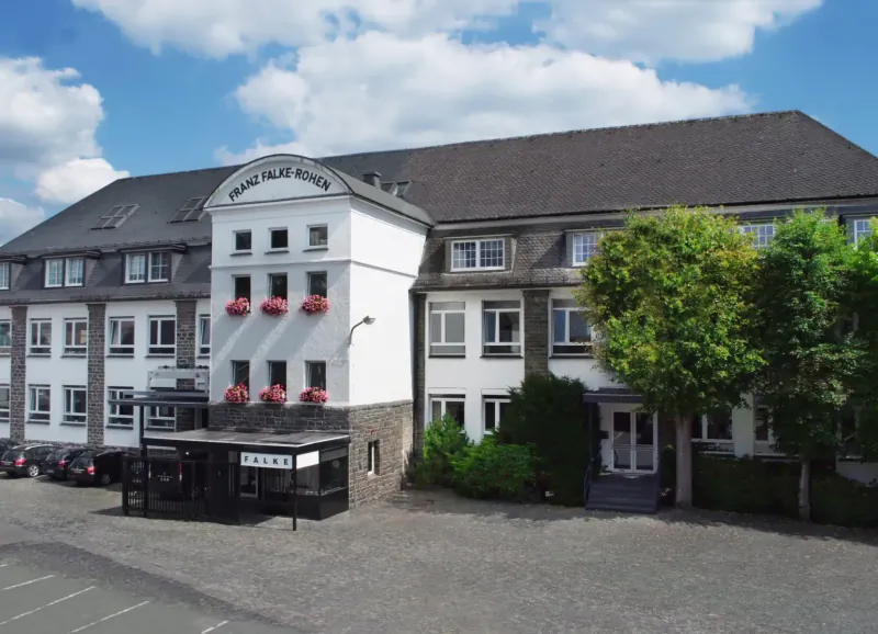 Falke Krahlaus building with 'FALKE KRAHLAUS' sign, flower boxes, trees, and cars in courtyard under blue sky.