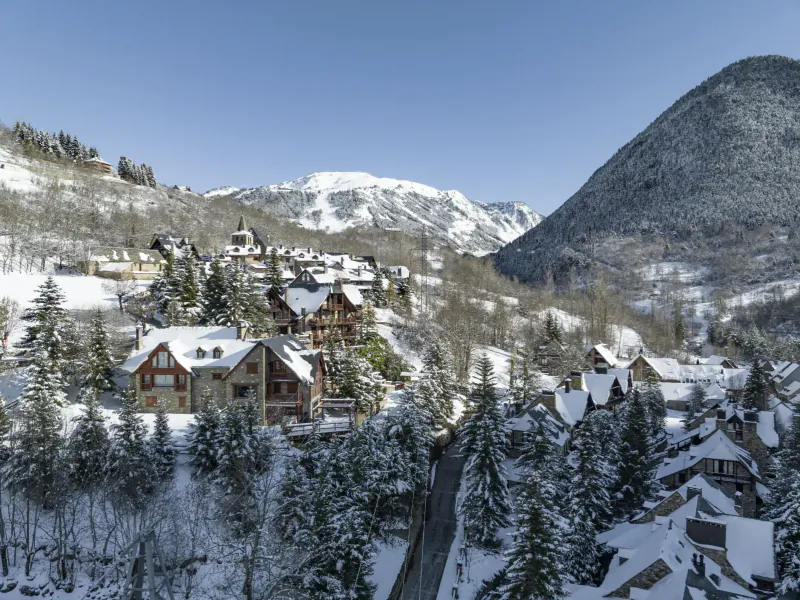 Snowy Val d'Aran village in Catalan Pyrenees with chalets, pine trees, and mountains under blue sky