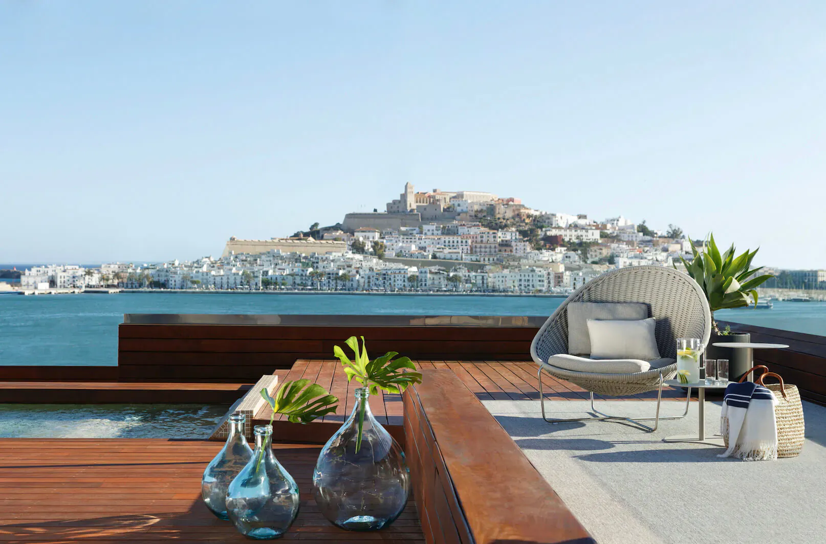 Rooftop terrace at Ibiza Gran Hotel with wicker chair, blue glass vases, plants, and sea view of white Ibizan town on hill