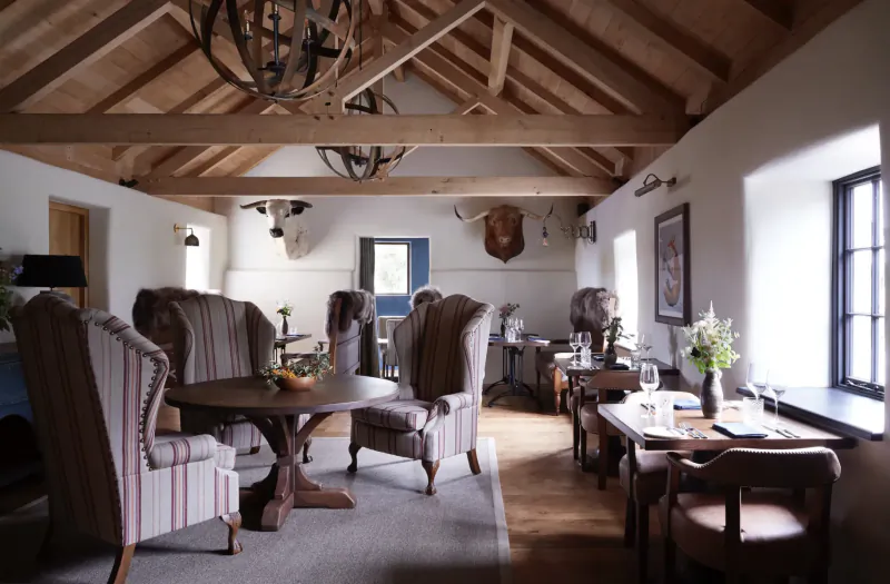 Cozy dining room in luxury retreat with wooden beams, mounted deer heads, armchairs, round table with fruit, and window views.