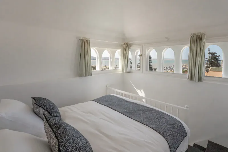 Bright white attic bedroom in Essaouira villa with arched windows overlooking sea, plush bed, and stairs.
