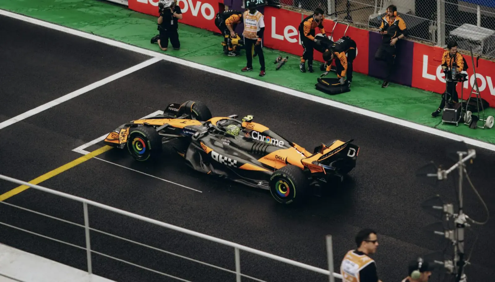 McLaren orange Formula 1 car on Le Mans pit lane with Lenovo branding and crew nearby