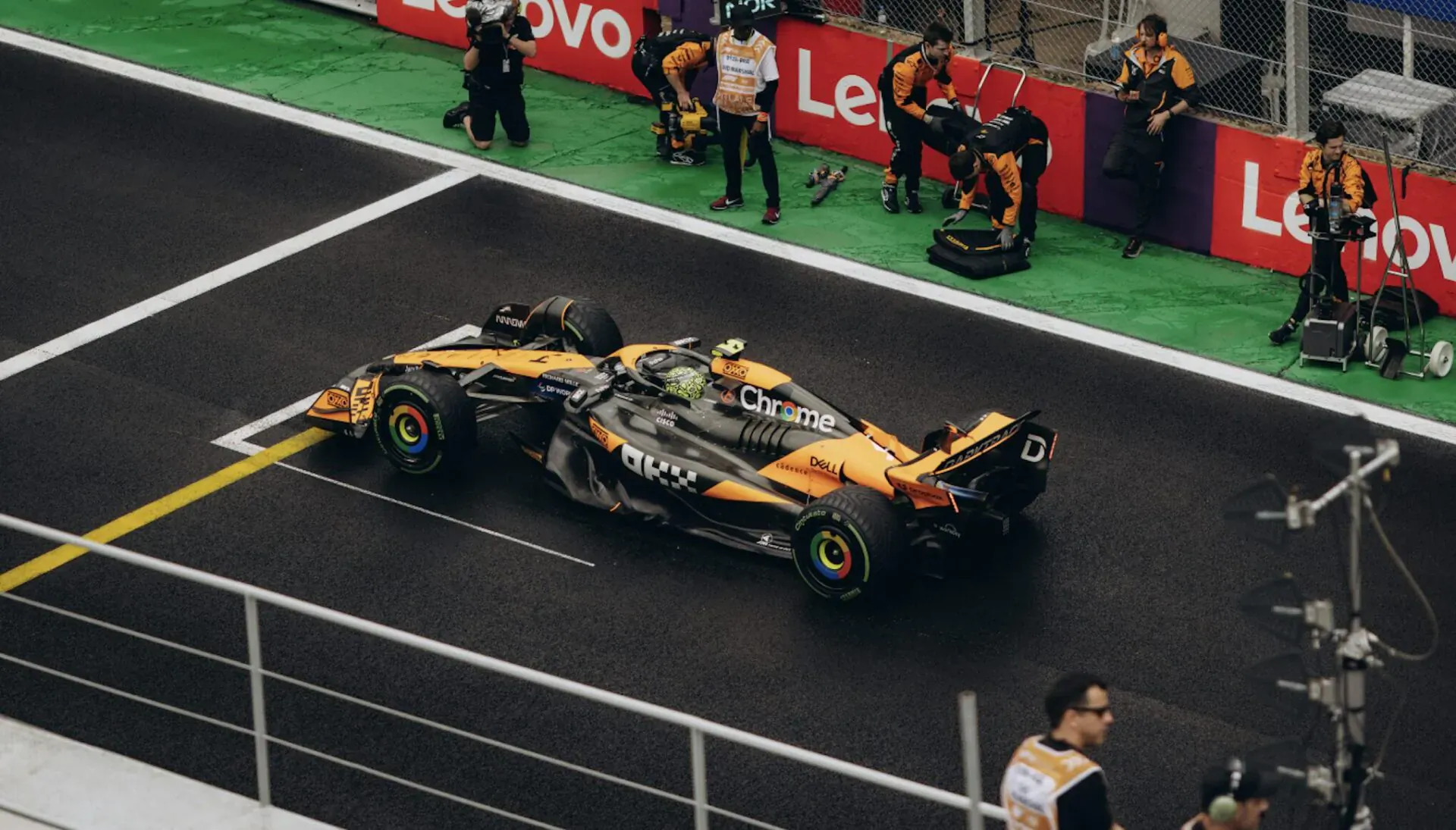 McLaren orange Formula 1 car on Le Mans pit lane with Lenovo branding and crew nearby
