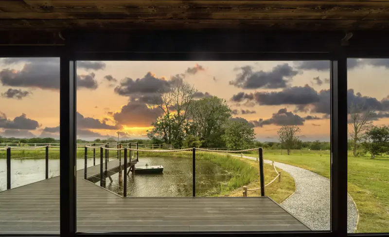 Luxury glamping pod window framing sunset over lake, dock with boat, and wooded path in West Wales.