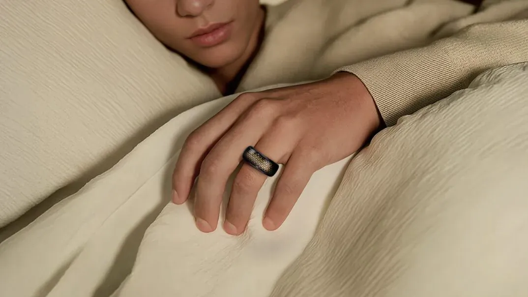 Close-up of hand wearing black VERTU AI Meta Ring, resting on beige bedding in bed