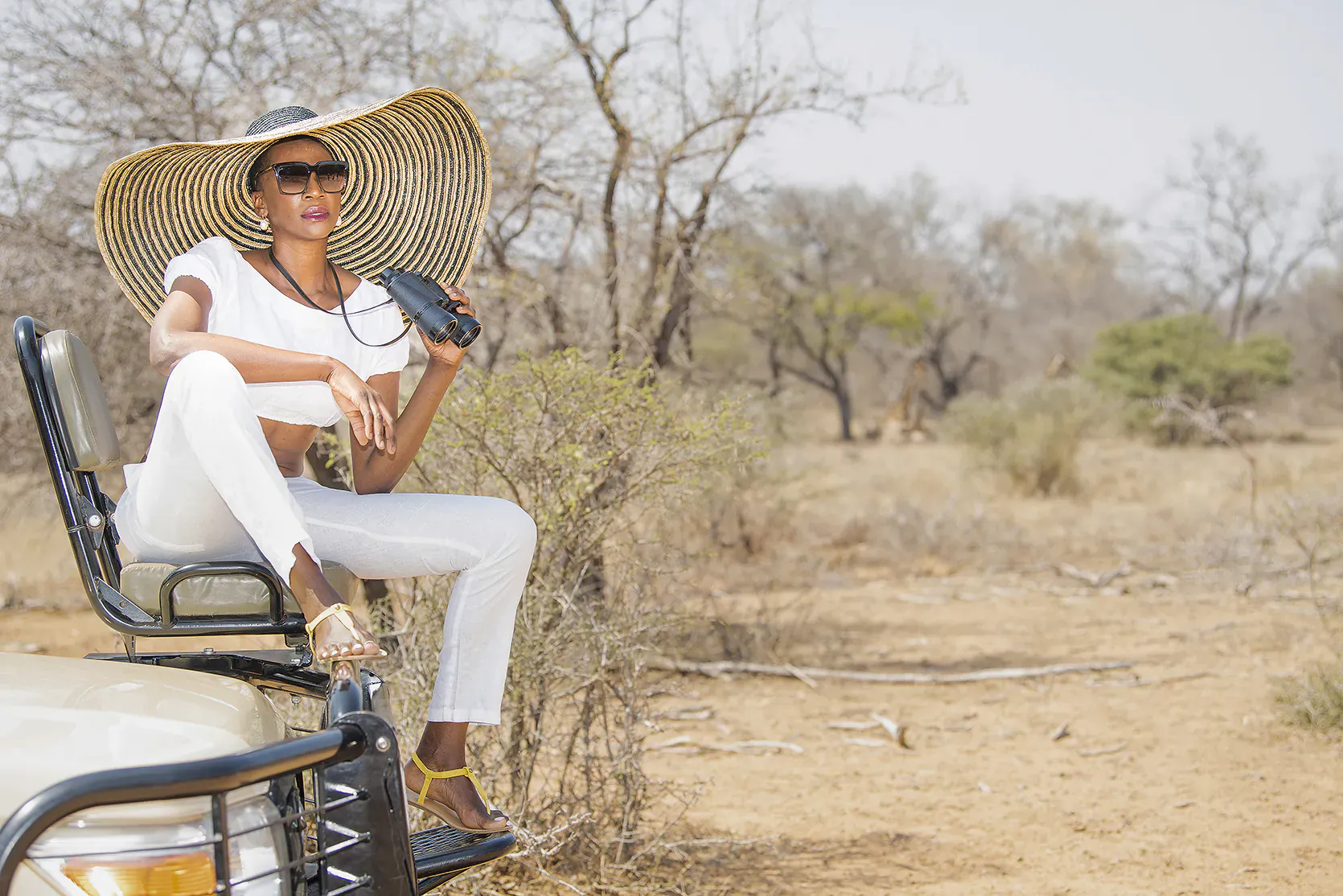 Woman in white outfit and wide-brim hat sits on safari jeep in dry South African bushveld, sunglasses on, holding handset