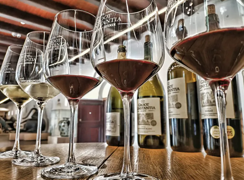 Row of wine glasses with white and red wines from Groot Constantia bottles on wooden bar, winery interior.