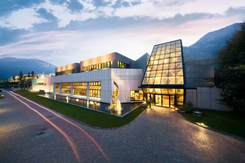 Modern Ferrari museum with glass pyramid entrance, giraffe sculpture, and mountain backdrop at dusk