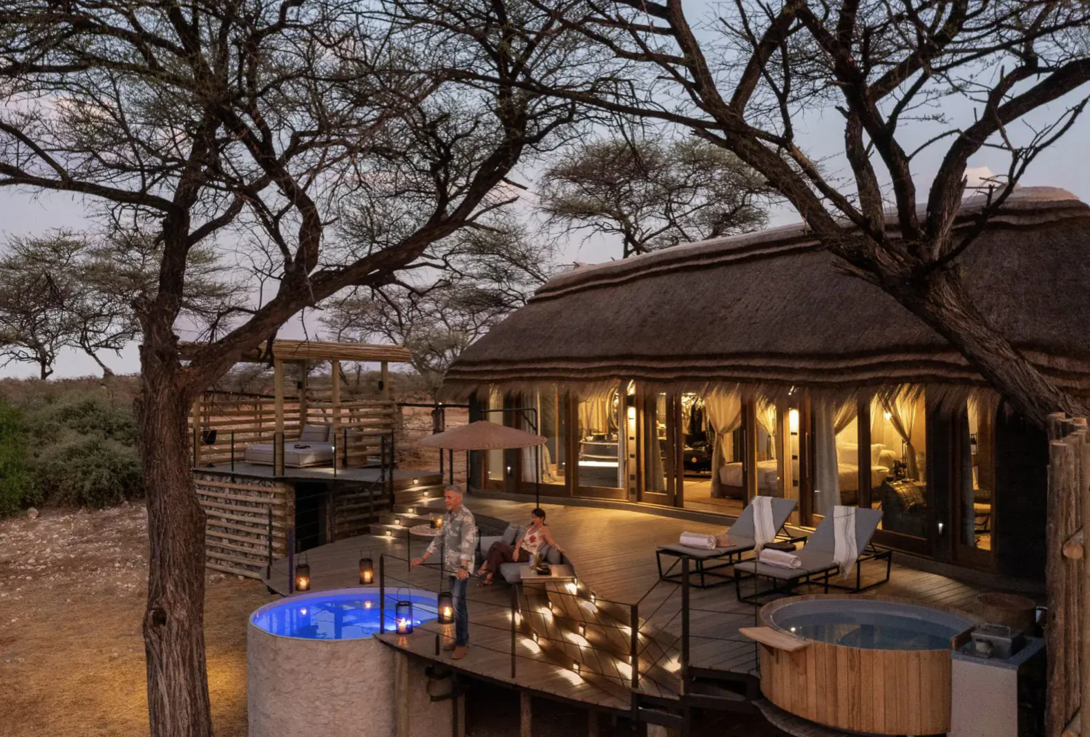 Luxury Camp Kala lodge in Etosha savanna at dusk, with glowing blue hot tub, lounge chairs, people relaxing on deck under acacia trees.