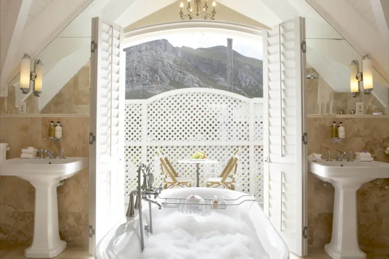 Luxurious bathroom with clawfoot tub full of bubbles, open shutters to mountain view, balcony table, sinks