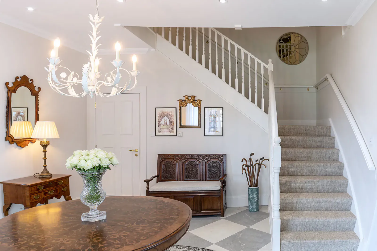 Elegant luxury manor home entrance with chandelier, wooden bench, flowers, and carpeted staircase.